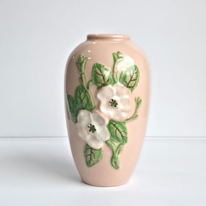 May include: A pink ceramic vase with a floral design. The vase features embossed white flowers with green leaves and stems. The vase has a smooth, glossy finish and a rounded shape, suitable for home decor.