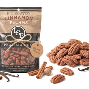 May include: A stand-up pouch of Hill Country Cinnamon Pecans with a clear window, tied with twine and a round tag reading Made in Texas. The bag displays the text Cinnamon Pecans, Lone Star Provisioning, and Net Wt. 8 oz (227g). Loose pecans, cinnamon sticks, and vanilla beans surround the bag.