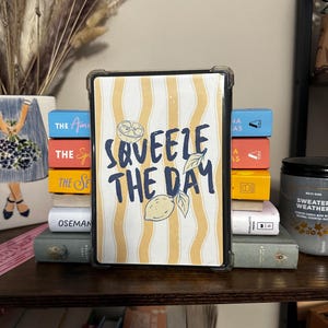 May include: A tablet case with a yellow and white striped design and the words "Squeeze the Day" in navy blue. The case is propped up against a stack of books on a wooden shelf.