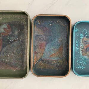 May include: Three rectangular decorative trays with rounded corners. Each tray features a painted portrait of a person. The trays are in shades of green, blue, and brown. The artwork has a vintage, distressed appearance.