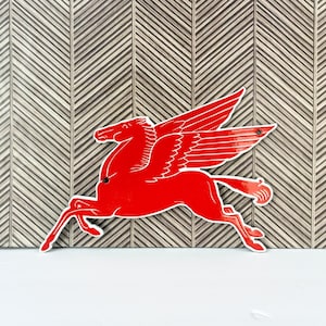 Könnte beinhalten: Ein leuchtend rotes Metallschild in Form eines Pegasus, dem mythischen geflügelten Pferd. Die Silhouette zeigt ein Pferd in einer dynamischen Laufpose mit großen, ausgebreiteten Flügeln. Zwei kleine kreisförmige Löcher sind am Körper sichtbar.