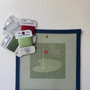 May include: Needlepoint canvas featuring a golf course scene with a green, flag, and golf ball. The canvas is framed in navy blue. Spools of red, green, and silver thread are arranged around the canvas, with labels reading "Tilli Tomas essentials" and "Kreinik".