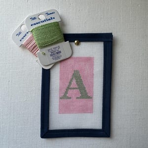 May include: A needlepoint project featuring a pink square with a green letter "A" inside a navy blue frame. Two spools of embroidery floss in pink and green are placed above the frame. The spools have the text "Tilli Tomas essentials".