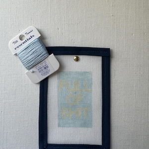 May include: A needlepoint canvas with the text "FULL OF SHIT" in yellow, framed in navy blue. A spool of light blue thread is next to the canvas. The canvas is attached to a white mesh.