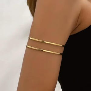 May include: Gold-colored arm cuff jewelry. The image shows two thin, parallel bands of gold metal worn on the upper arm. The jewelry is simple and elegant, reflecting a minimalist design. The background is a neutral color, highlighting the arm cuffs.