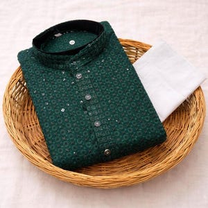 May include: A dark green kurta shirt with detailed embroidery and silver embellishments, accompanied by white trousers. The shirt has a mandarin collar and button-down front. The outfit is displayed in a woven basket.