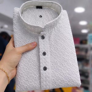 Woman holding a folded embroidered men’s kurta with mandarin collar and textured floral design, displayed in store.
