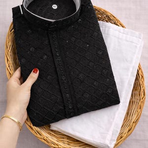 K&ouml;nnte beinhalten: Schwarzes besticktes Kurta-Hemd mit Stehkragen, kombiniert mit einer wei&szlig;en Hose. Das Hemd weist aufwendige Details auf und ist ordentlich in einem Weidenkorb gefaltet. Ein Gr&ouml;&szlig;enetikett 40 ist sichtbar.