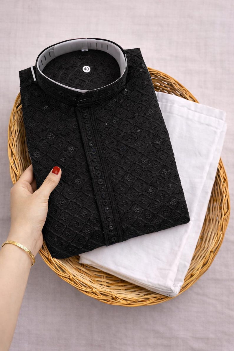 K&ouml;nnte beinhalten: Schwarzes besticktes Kurta-Hemd mit Stehkragen, kombiniert mit einer wei&szlig;en Hose. Das Hemd weist aufwendige Details auf und ist ordentlich in einem Weidenkorb gefaltet. Ein Gr&ouml;&szlig;enetikett 40 ist sichtbar.