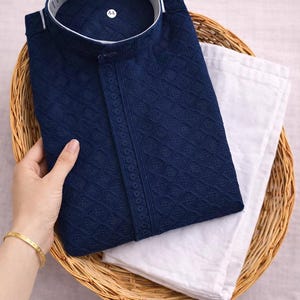 K&ouml;nnte beinhalten: Ein marineblaues, besticktes Kurta-Hemd mit Stehkragen, kombiniert mit einer wei&szlig;en Hose. Das Hemd weist aufwendige Rautenmuster auf und ist ordentlich in einem Weidenkorb gefaltet. Ein goldenes Armband ist sichtbar.