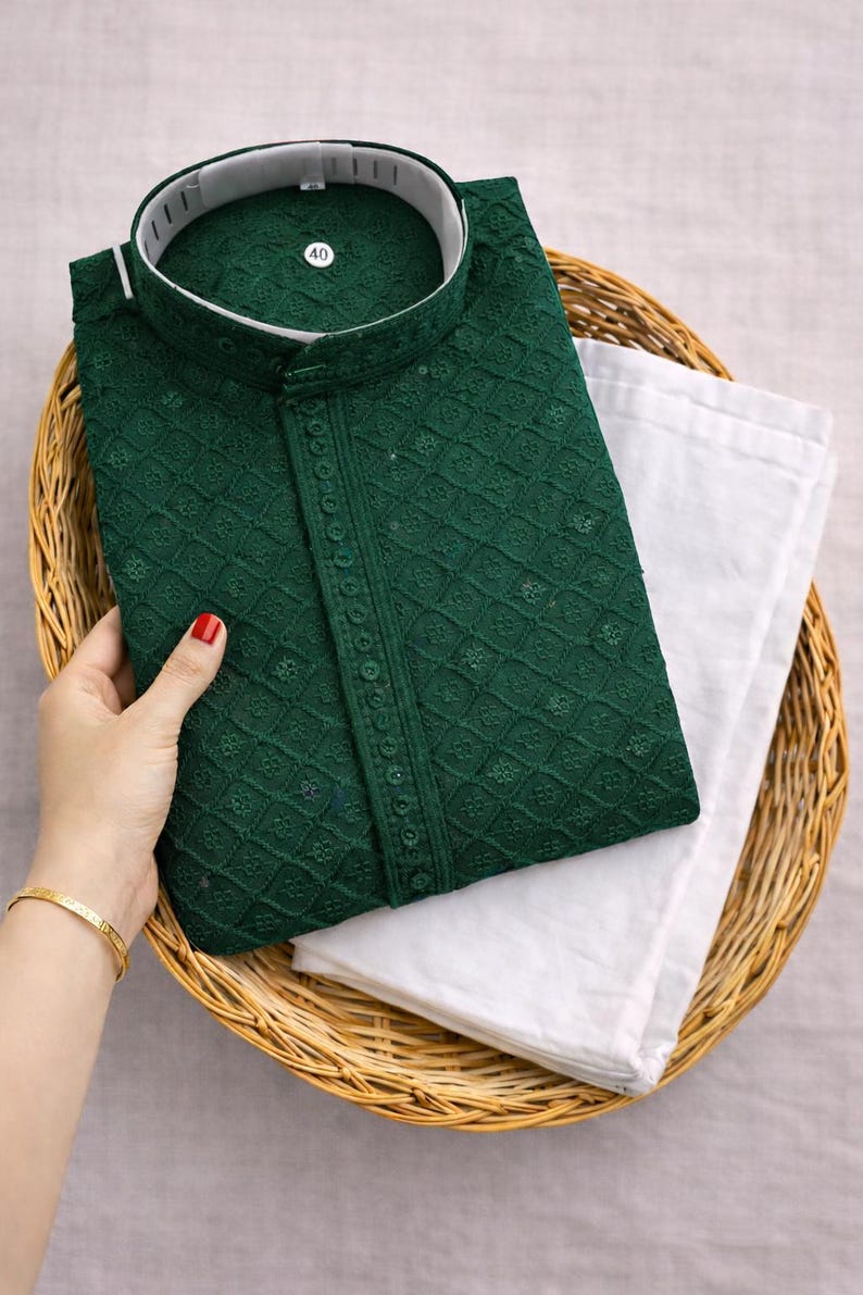 K&ouml;nnte beinhalten: Ein dunkelgr&uuml;nes, besticktes Kurta mit wei&szlig;em Kragen und einer passenden wei&szlig;en Hose, pr&auml;sentiert in einem Weidenkorb. Das Kurta weist aufwendige Details und eine Knopfleiste auf. Das Gr&ouml;&szlig;enetikett lautet 40.
