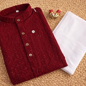 K&ouml;nnte beinhalten: Ein bordeauxfarbener Kurta mit goldenen Kn&ouml;pfen und Stehkragen, kombiniert mit einer wei&szlig;en Hose. Der strukturierte Stoff des Kurtas ist sichtbar. Dekorative Figuren sind in der N&auml;he der Kleidung platziert. Das Outfit wird auf einer gewebten Oberfl&auml;che pr&auml;sentiert.