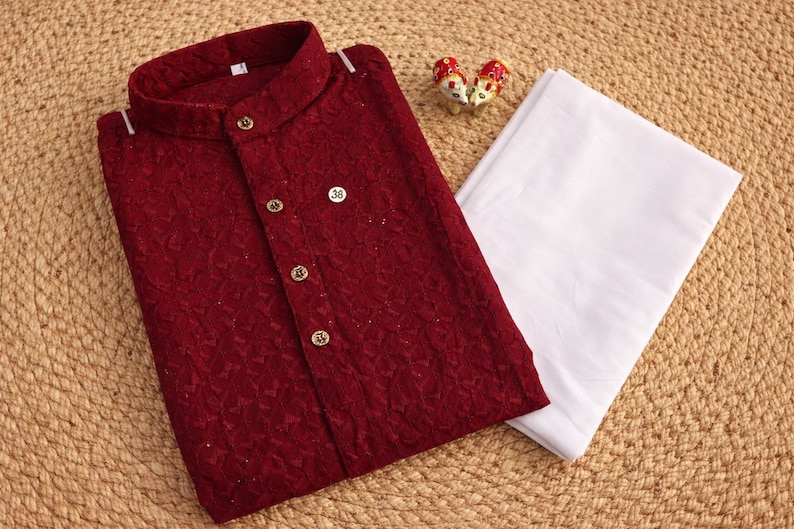 K&ouml;nnte beinhalten: Ein bordeauxfarbener Kurta mit goldenen Kn&ouml;pfen und Stehkragen, kombiniert mit einer wei&szlig;en Hose. Der strukturierte Stoff des Kurtas ist sichtbar. Dekorative Figuren sind in der N&auml;he der Kleidung platziert. Das Outfit wird auf einer gewebten Oberfl&auml;che pr&auml;sentiert.