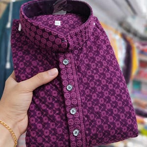 Woman holding a folded embroidered men’s kurta with mandarin collar and textured floral design, displayed in store.