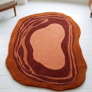 May include: A unique, abstract rug with a layered design in shades of orange and brown. The rug has an irregular, organic shape, adding a modern touch to any room. The rug's dimensions are approximately 60 inches by 48 inches.