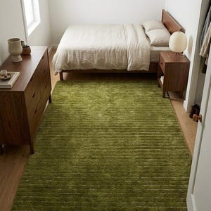 May include: A bedroom scene featuring a green shag rug, a wooden bed frame with beige bedding, and a matching nightstand. A wooden dresser is on the left. The room has a minimalist design with natural light from a window.