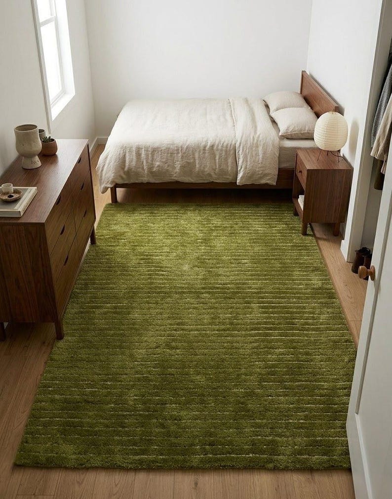 May include: A bedroom scene featuring a green shag rug, a wooden bed frame with beige bedding, and a matching nightstand. A wooden dresser is on the left. The room has a minimalist design with natural light from a window.