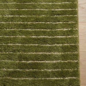 May include: A close-up of a green rug with horizontal white stripes. The rug has a shaggy texture and is a shade of olive green. The rug is on a light brown wooden floor.
