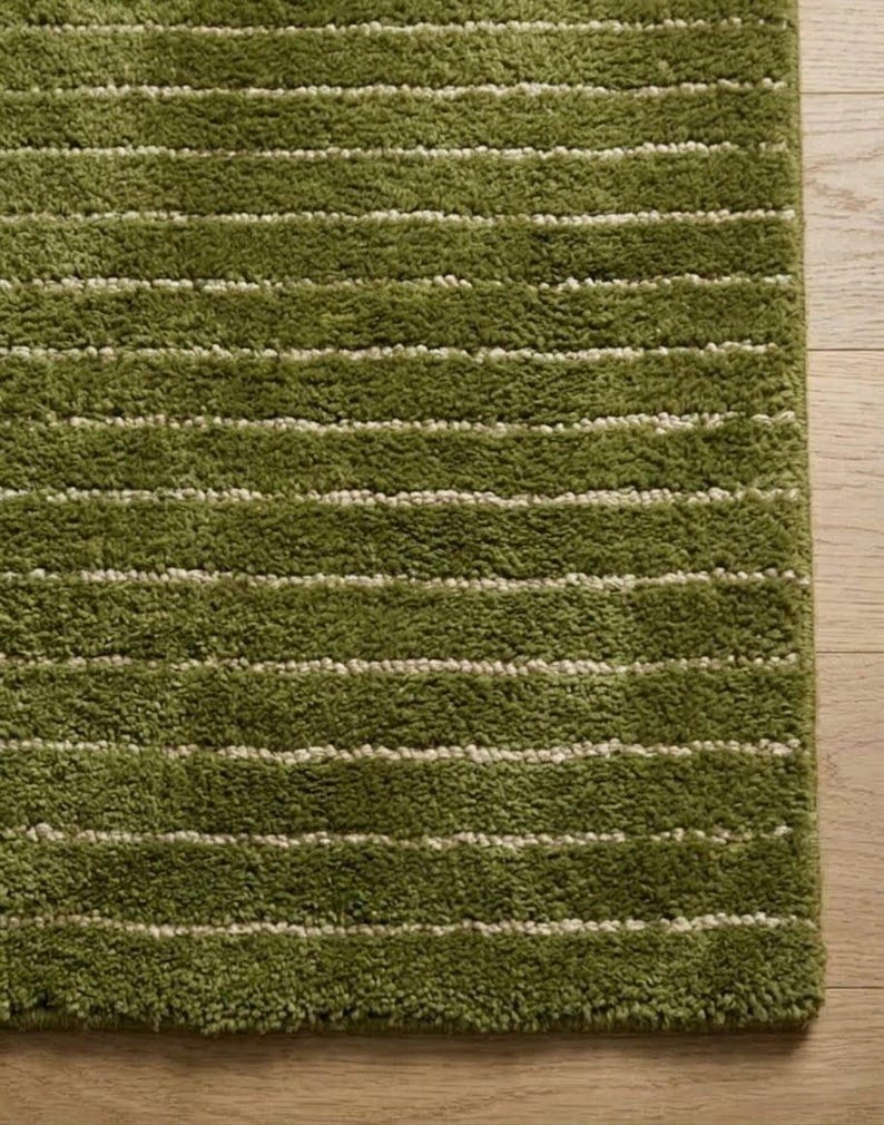May include: A close-up of a green rug with horizontal white stripes. The rug has a shaggy texture and is a shade of olive green. The rug is on a light brown wooden floor.
