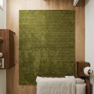 May include: Overhead view of a bedroom with a green rectangular rug with horizontal white stripes. The rug is centered on a light wood floor. A bed with a beige comforter and pillows is in the lower right corner. A wooden nightstand and cabinet are also visible.