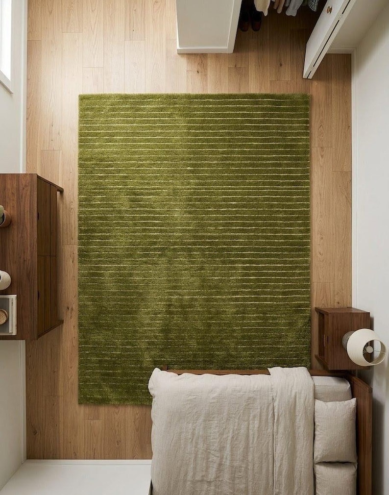 May include: Overhead view of a bedroom with a green rectangular rug with horizontal white stripes. The rug is centered on a light wood floor. A bed with a beige comforter and pillows is in the lower right corner. A wooden nightstand and cabinet are also visible.
