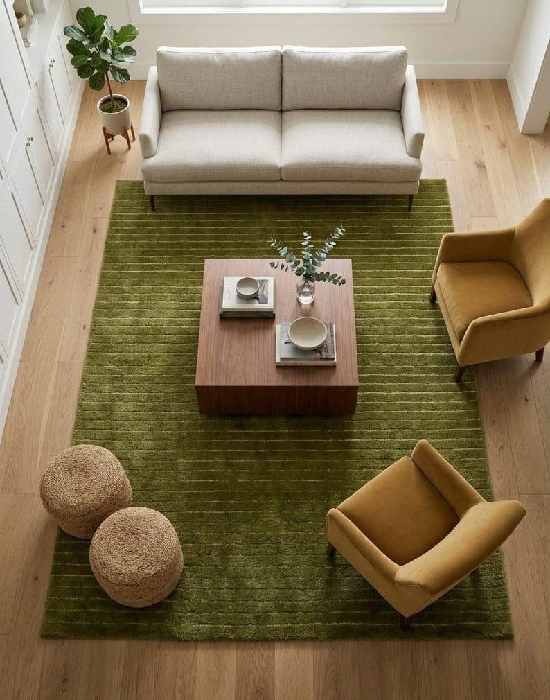 May include: An overhead shot of a living room with a light-colored sofa, two mustard-yellow armchairs, and a square wooden coffee table on a green rug. Two woven poufs and a potted plant are also visible.