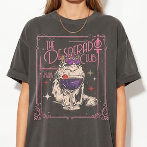 May include: Dark gray t-shirt featuring a whimsical illustration. The graphic includes a cat wearing sunglasses and holding a cocktail, with the text "The Desperado Club" and "So Fun It Hurts" in a pink frame.