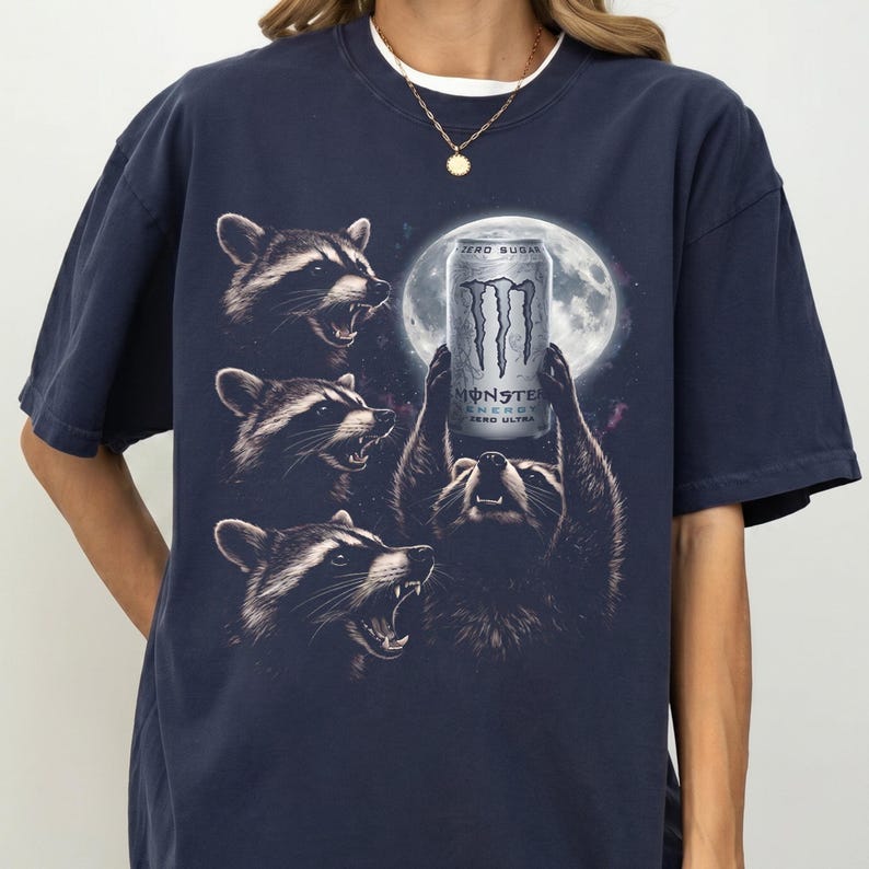 May include: Navy blue t-shirt featuring a graphic of raccoons surrounding a Monster Energy drink can in front of a full moon. The can reads "ZERO SUGAR" and "MONSTER ENERGY ZERO ULTRA". The raccoons are detailed with black and white fur.