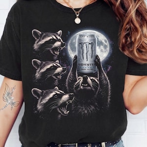 May include: Black t-shirt featuring a graphic of raccoons with a full moon in the background. One raccoon holds a Monster Energy drink can that reads "ZERO SUGAR" and "ZERO ULTRA". The design is detailed and eye-catching.