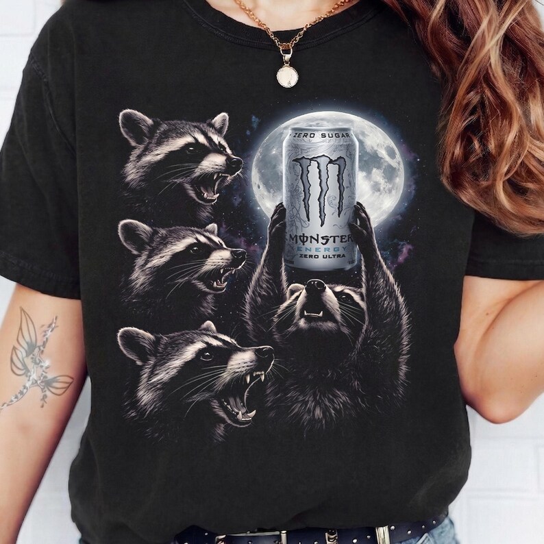 May include: Black t-shirt featuring a graphic of raccoons with a full moon in the background. One raccoon holds a Monster Energy drink can that reads "ZERO SUGAR" and "ZERO ULTRA". The design is detailed and eye-catching.