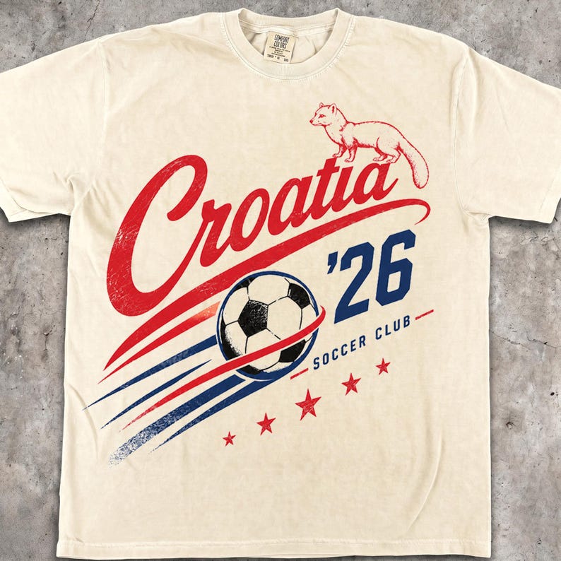 May include: A cream-coloured short-sleeve t-shirt featuring a graphic print with the red text Croatia, a small red marten illustration, a black and white football, the blue text '26 SOCCER CLUB, and five red stars arranged in a diagonal line.