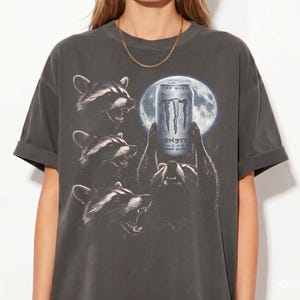 May include: Dark gray t-shirt featuring a graphic of raccoons holding a Monster Energy drink can in front of a full moon. The shirt has a vintage, worn look, with rolled-up sleeves.