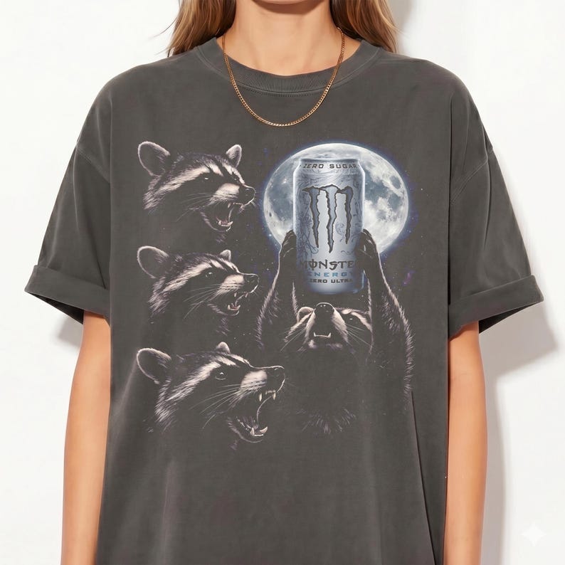May include: Dark gray t-shirt featuring a graphic of raccoons holding a Monster Energy drink can in front of a full moon. The shirt has a vintage, worn look, with rolled-up sleeves.