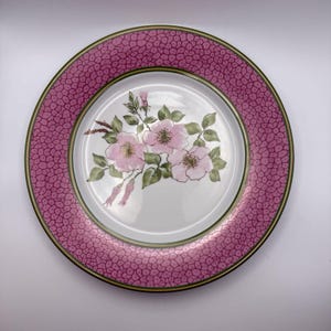 Peut inclure: Une assiette décorative ronde avec un centre blanc orné d'un motif floral de fleurs roses et de feuilles vertes. Le bord est rose texturé avec une bordure vert foncé.