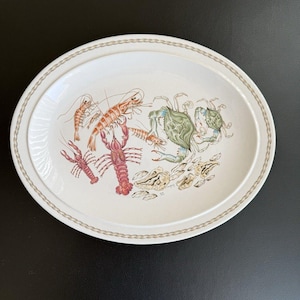 Puede incluir: Plato ovalado para servir con fondo color crema y borde con diseño de cadena repetida. El plato presenta ilustraciones de varios mariscos, incluyendo camarones, langostas y cangrejos, en tonos de rojo, naranja, verde y azul.