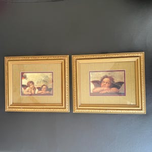 May include: Two framed prints featuring cherubic figures. Each print has a gold-colored ornate frame with a gold-colored mat. The prints depict cherubs with wings, resting their chins on their hands, with a soft, classical style.