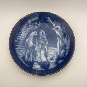 Może przedstawiać: Okrągły, niebiesko-biały talerz dekoracyjny przedstawiający scenę dwóch postaci w szatach pod drzewem. Talerz ma ciemnoniebieską obwódkę, a na dole wydrukowano rok "1972".
