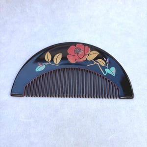 Puede incluir: Un peine de pelo negro semicircular con un diseño floral. El peine presenta una flor roja con hojas doradas y verdes. Los dientes del peine son de color marrón oscuro. El peine probablemente está hecho de un material brillante.