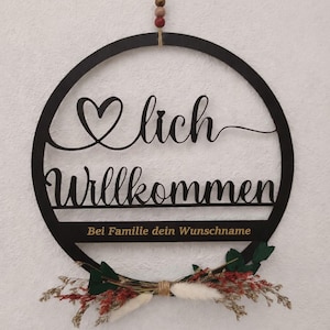 Könnte beinhalten: Schwarzes Holz-Willkommensschild mit einem Herz über dem Wort "Willkommen". Das Schild hat eine kreisförmige Form und ist mit Trockenblumen und Grün geschmückt. Der Text "Bei Familie dein Wunschname" ist ebenfalls enthalten.
