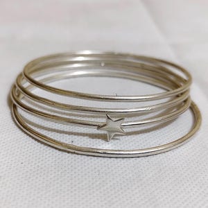 May include: A collection of silver bangles. One bangle has a small star-shaped charm. The thin, circular bangles are stacked together. The jewellery is presented on a white surface.
