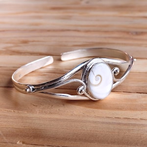 Puede incluir: Una pulsera de plata con un centro ovalado de concha blanca. La concha tiene un diseño en espiral. La pulsera tiene remolinos decorativos a cada lado de la concha.