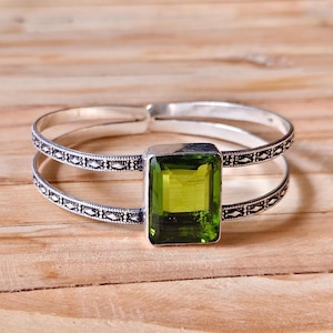 Può includere: Un bracciale a fascia in argento con design a doppio cerchio, un intricato motivo inciso e una grande pietra preziosa rettangolare sfaccettata color verde lime incastonata al centro. Il metallo ha una finitura lucida con accenti scuri.