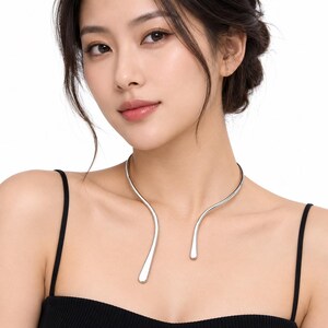 Op de afbeelding: Een zilverkleurige metalen choker-halsketting met een open, asymmetrisch ontwerp en twee gladde, taps toelopende uiteinden die sierlijk om de nek buigen. Het gepolijste, reflecterende oppervlak vormt een strakke, doorlopende metalen lijn.