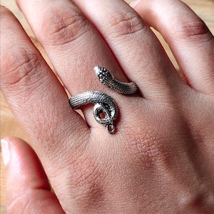 Puede incluir: Un anillo de metal plateado con forma de serpiente enroscada. El diseño presenta un patrón texturizado similar a escamas en todo el cuerpo de la serpiente, con la cabeza y la cola superpuestas para crear un estilo de banda abierta y ajustable.