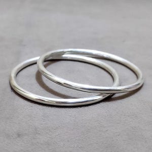 Peut inclure: Deux bracelets de couleur argentée, chacun avec un design lisse et arrondi. Les bracelets sont circulaires et semblent être en métal. Ils reposent sur une surface grise. L'image est bien éclairée, soulignant la qualité réfléchissante des bijoux.