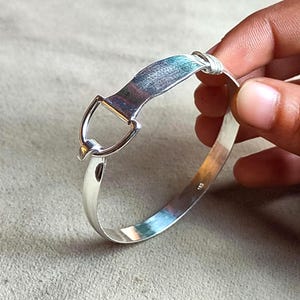 May include: A silver-colored bangle bracelet with a unique design. The bracelet features a rectangular plate connected to a D-shaped loop and a rounded band. The metal has a polished finish, reflecting light. The bracelet is held by a hand.