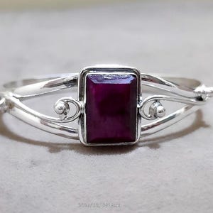 Può includere: Un bracciale in argento con una gemma rettangolare rosso scuro al centro. La gemma è incastonata in una cornice quadrata. Il bracciale ha un design delicato e curvo con volute decorative e piccole perle d'argento.