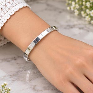 May include: A polished silver-tone bangle bracelet with a smooth, flat surface and a rounded profile. The bracelet features a clean, reflective metallic finish and a simple, continuous circular band design that rests comfortably on the wrist.