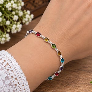 May include: A silver chain bracelet featuring a series of small, oval-shaped, multi-coloured gemstones in shades of red, yellow, green, blue, and purple. The stones are set in individual silver bezels connected by thin silver links in a repeating pattern.