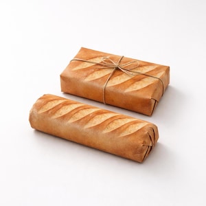 May include: Two fresh baguettes, one wrapped in brown paper and tied with twine, the other unwrapped. The baguettes have diagonal slashes on top, and the paper-wrapped loaf is rectangular. The background is white.
